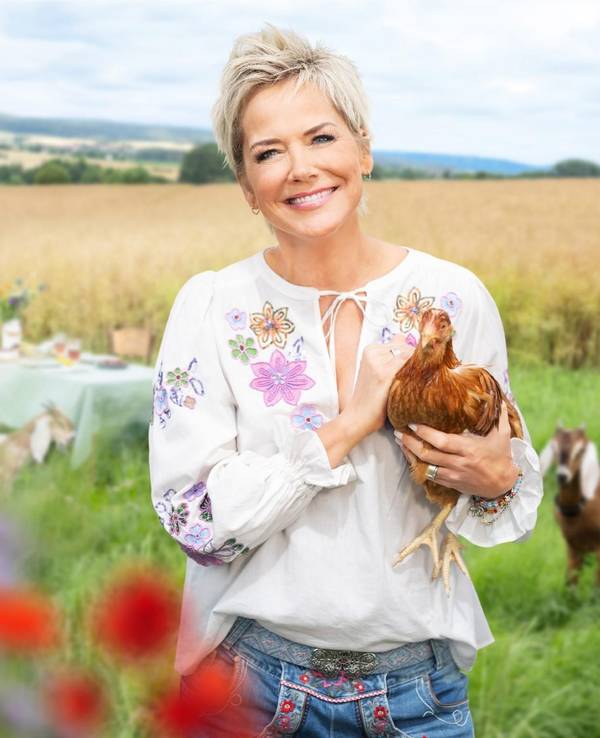 Moderatorin Inka Bause steht auf einer Wise und hält ein Huhn auf dem Arm. Im Hintergrund schaut eine Ziege.