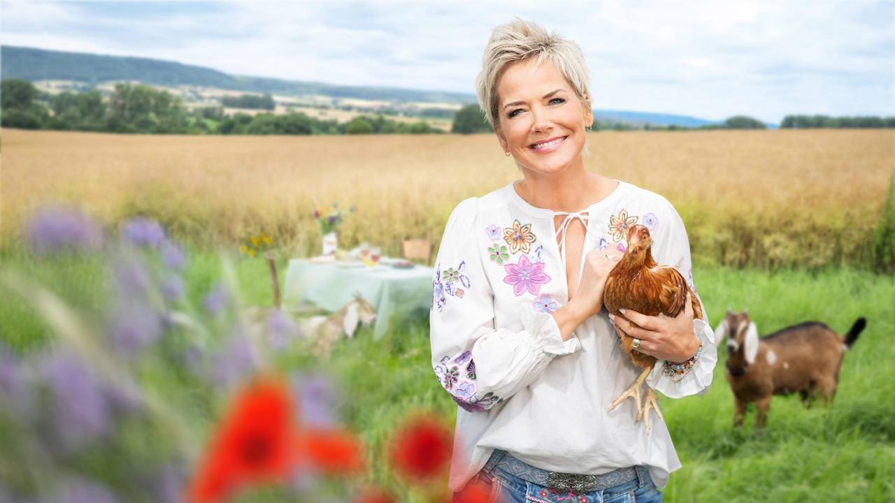 Moderatorin Inka Bause steht auf einer Wise und hält ein Huhn auf dem Arm. Im Hintergrund schaut eine Ziege.