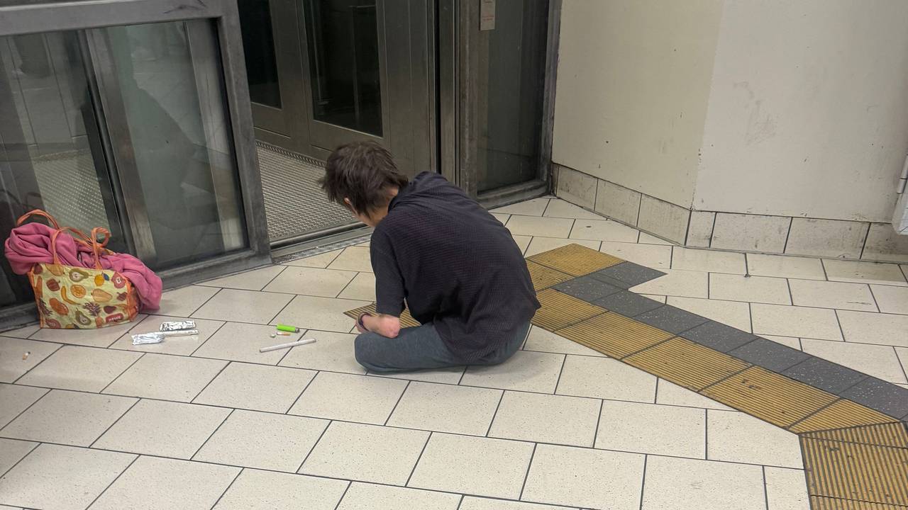 Ein Mann sitzt auf einem Fliesenboden, vor sich eine Spritze, ein Feuerzeug und andere Dinge.