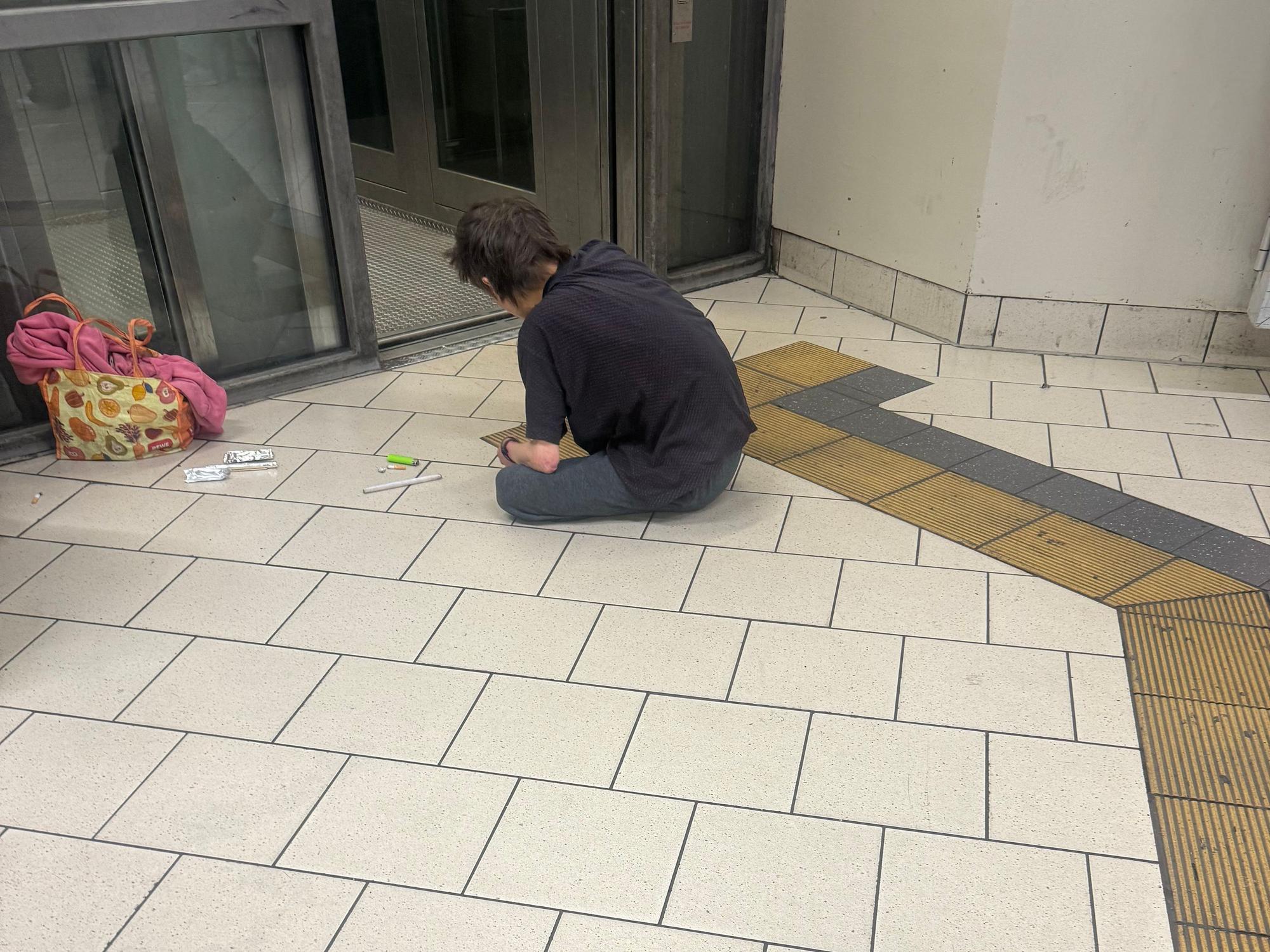 Ein Mann sitzt auf einem Fliesenboden, vor sich eine Spritze, ein Feuerzeug und andere Dinge.