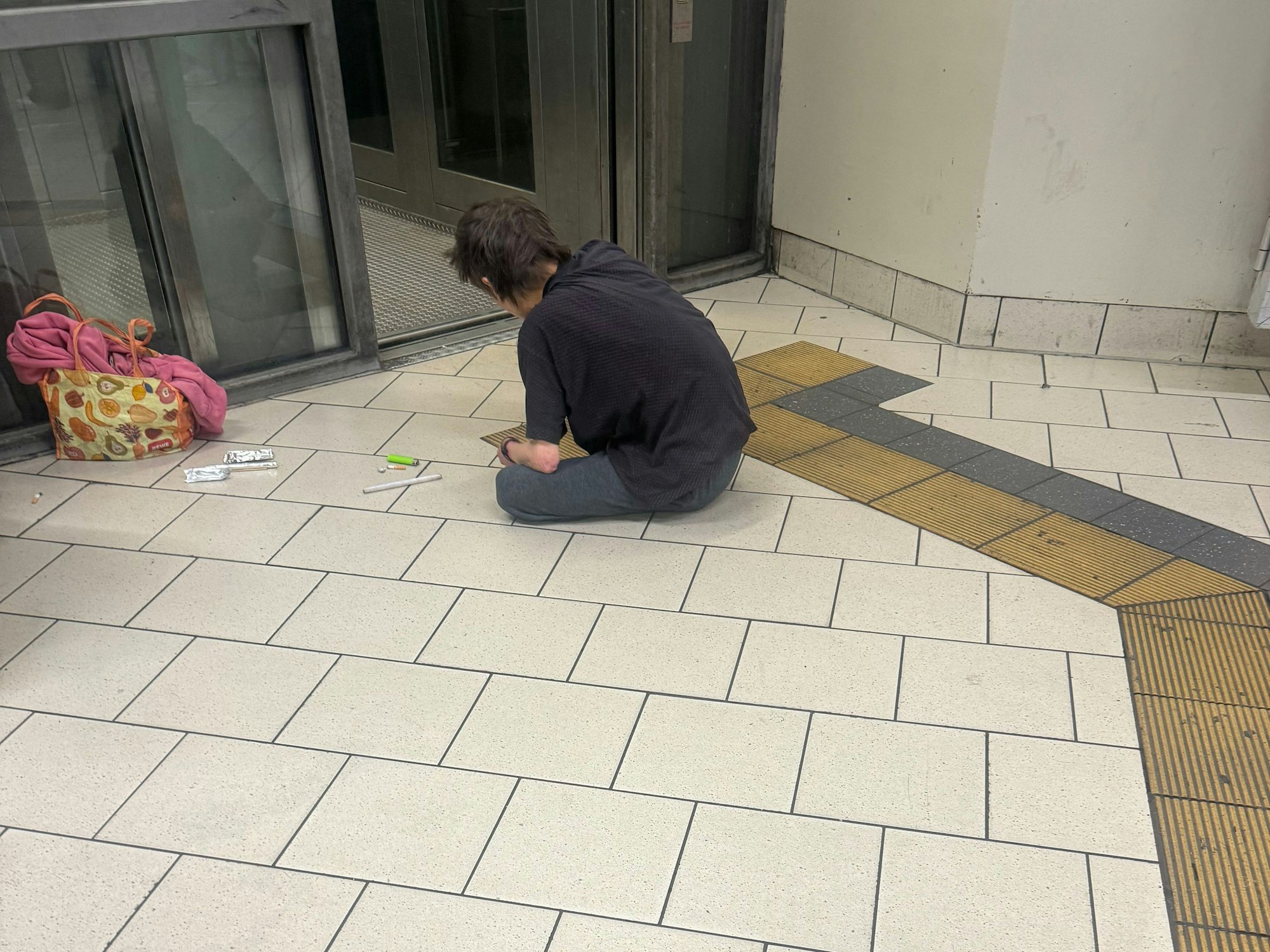 Ein Mann sitzt auf einem Fliesenboden, vor sich eine Spritze, ein Feuerzeug und andere Dinge.