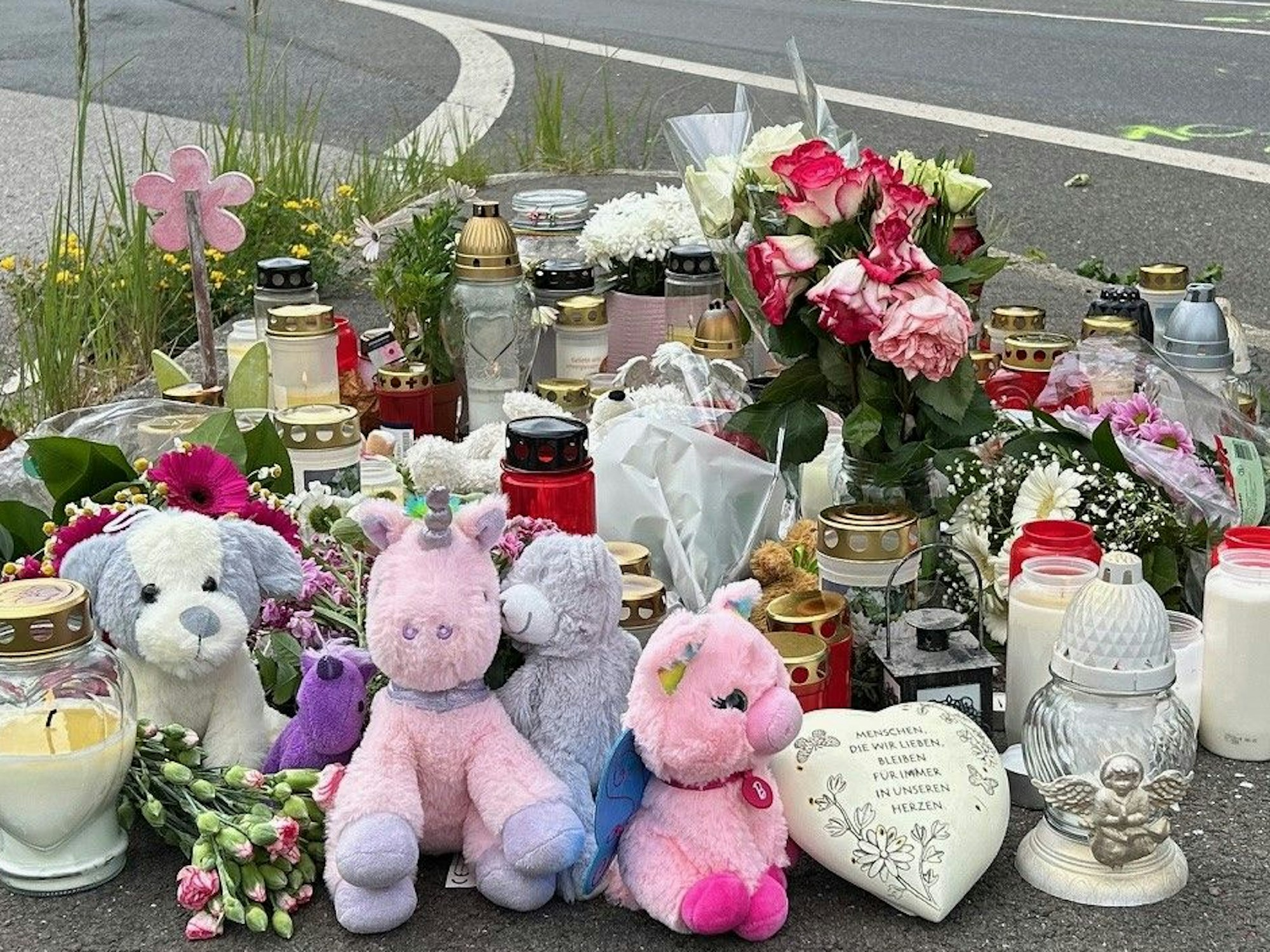 An einer Fahrbahn stehen Kerzen und Kuscheltiere, auch Blumen wurden niedergelegt.