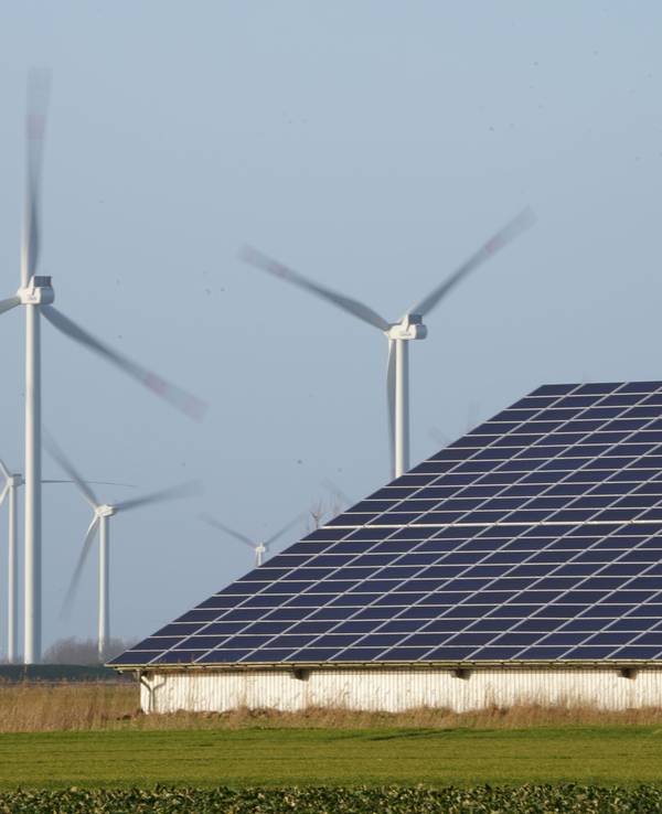 Windenergieanlagen stehen neben einer Halle mit Photovoltaik-Anlagen auf dem Dach.