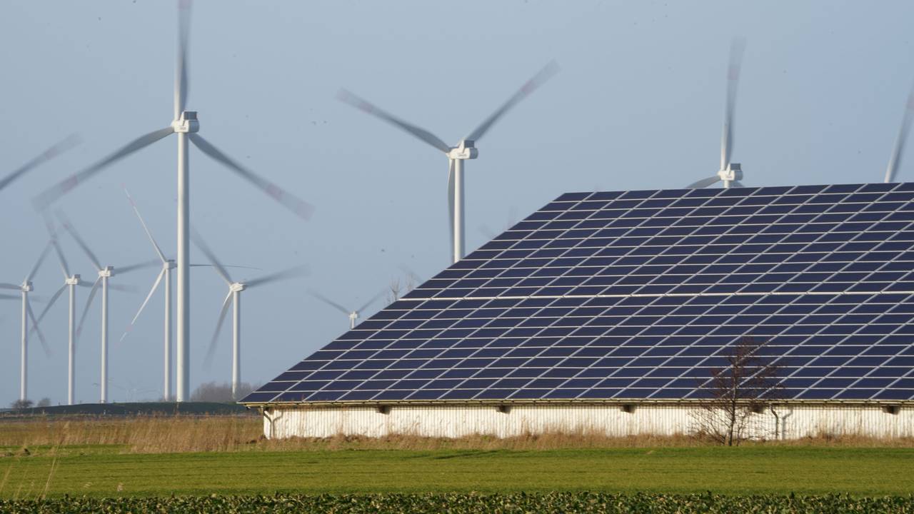 Windenergieanlagen stehen neben einer Halle mit Photovoltaik-Anlagen auf dem Dach.