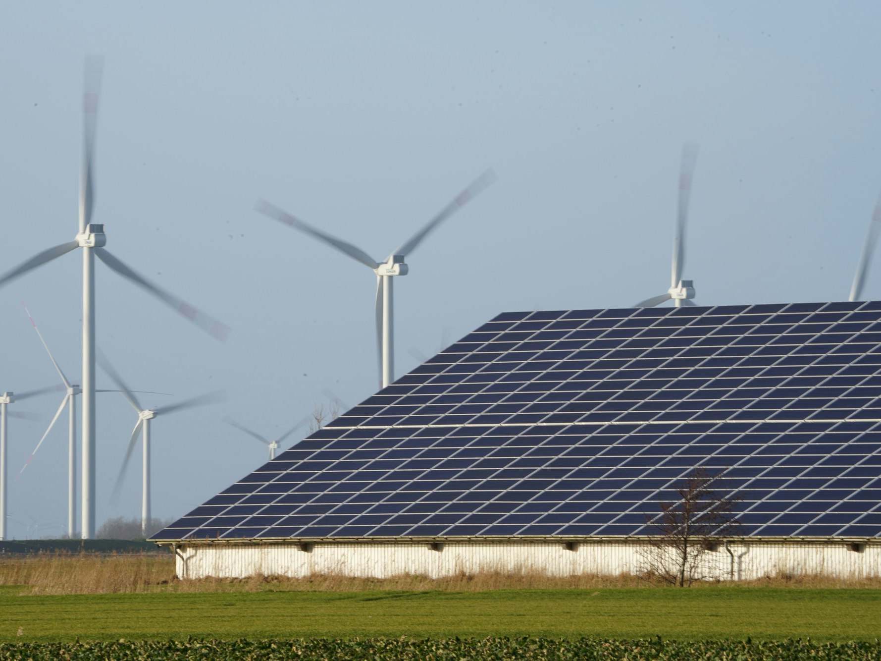Windenergieanlagen stehen neben einer Halle mit Photovoltaik-Anlagen auf dem Dach.