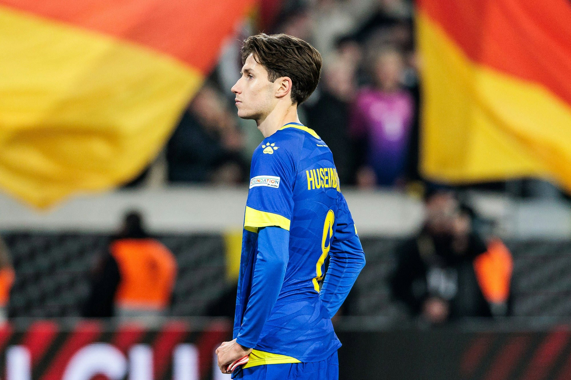 Denis Huseinbasic steht im Nationalmannschaftstrikot enttäuscht auf dem Rasen, im Hintergrund jubeln deutsche Fans.