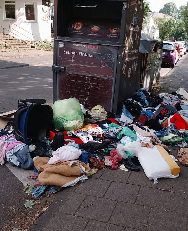 Um einen Altkleidercontainer liegen Kleidungs- und Wäschestücke auf dem Boden.