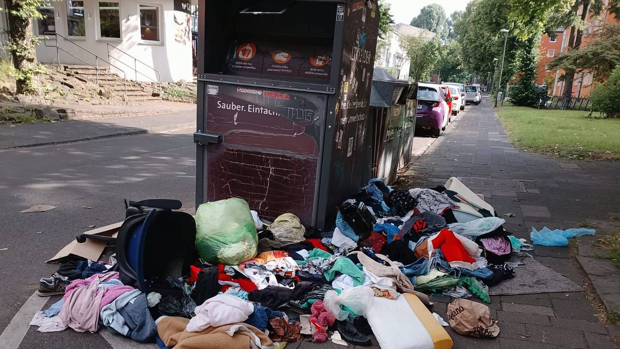 Um einen Altkleidercontainer liegen Kleidungs- und Wäschestücke auf dem Boden.