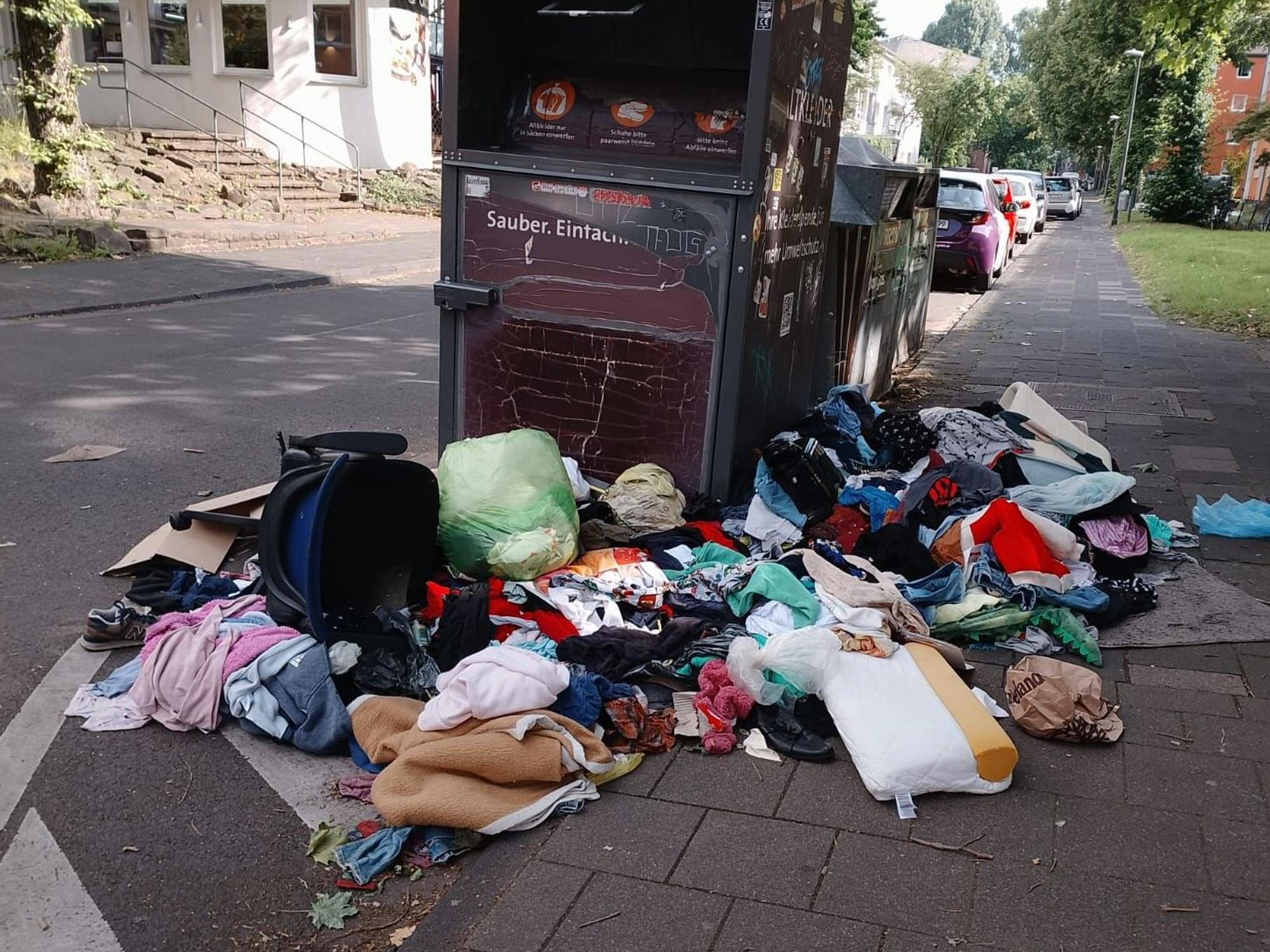 Um einen Altkleidercontainer liegen Kleidungs- und Wäschestücke auf dem Boden.