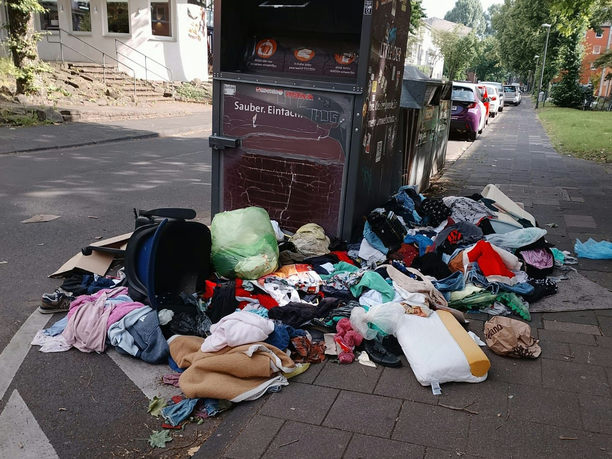 Um einen Altkleidercontainer liegen Kleidungs- und Wäschestücke auf dem Boden.