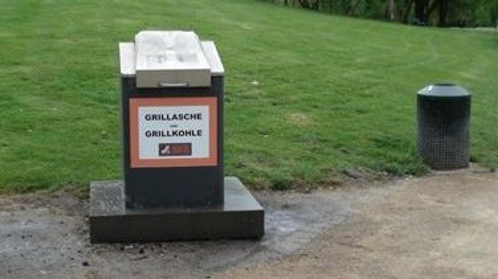 Ein kleiner Müllcontainer mit der Aufschrift Grillasche und Grillkohle steht am Rand einer Grünfläche.