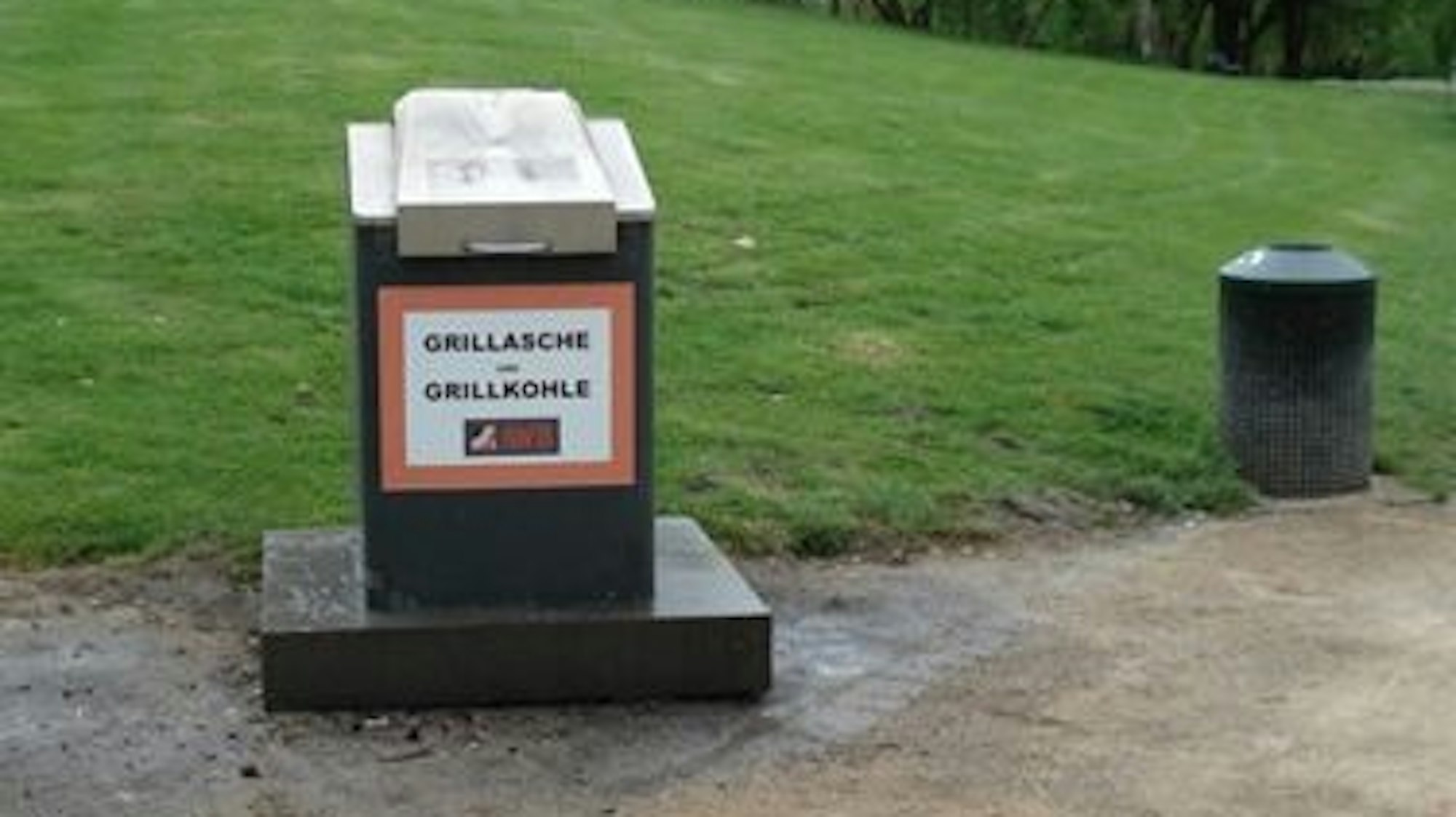 Ein kleiner Müllcontainer mit der Aufschrift Grillasche und Grillkohle steht am Rand einer Grünfläche.
