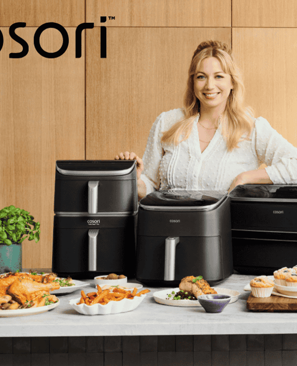 TV-Köchin Zora Klipp lächelnd hinter drei verschiedenen Cosori Heißluftfritteusen, umgeben von frisch zubereiteten Gerichten wie Hähnchen, Pommes und Muffins.
