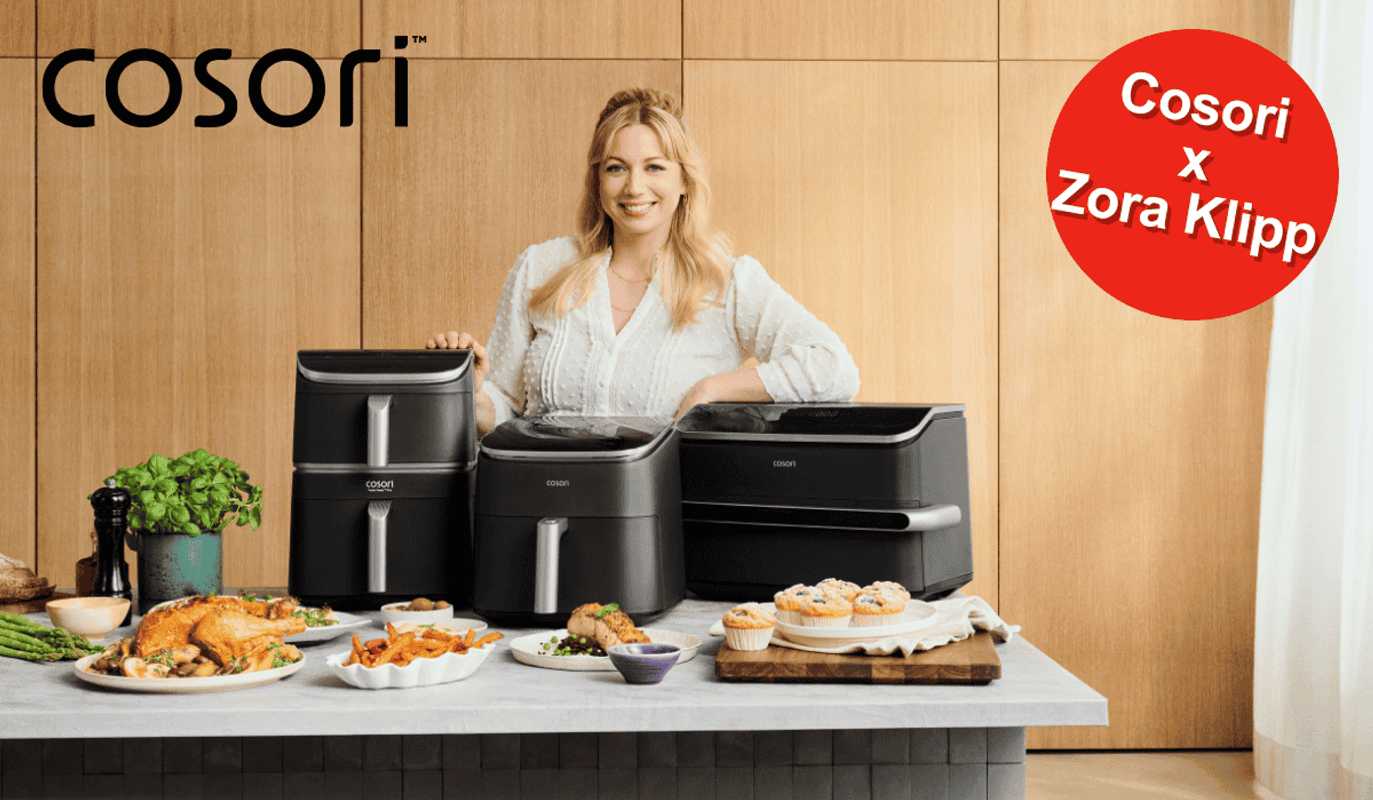 TV-Köchin Zora Klipp lächelnd hinter drei verschiedenen Cosori Heißluftfritteusen, umgeben von frisch zubereiteten Gerichten wie Hähnchen, Pommes und Muffins.