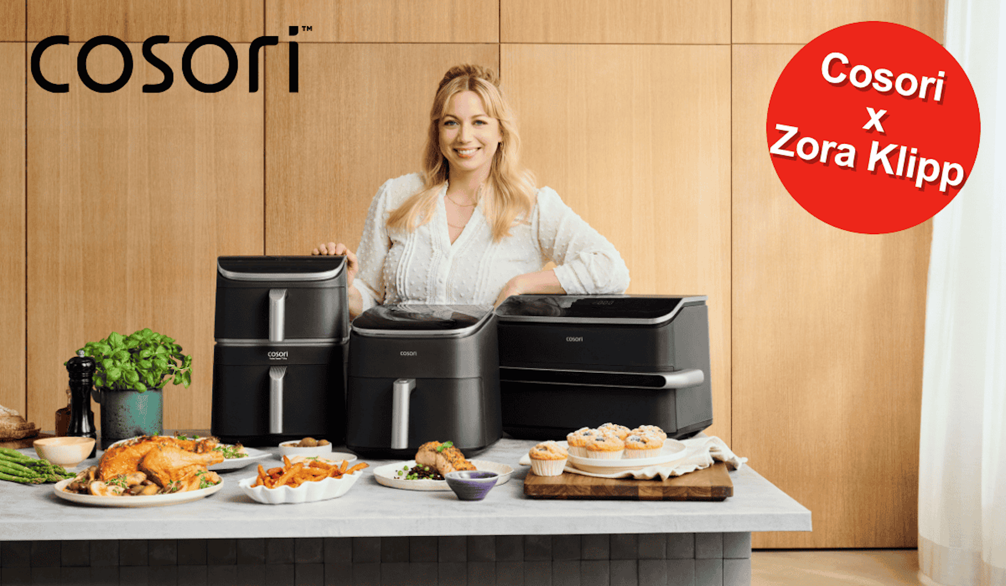TV-Köchin Zora Klipp lächelnd hinter drei verschiedenen Cosori Heißluftfritteusen, umgeben von frisch zubereiteten Gerichten wie Hähnchen, Pommes und Muffins.