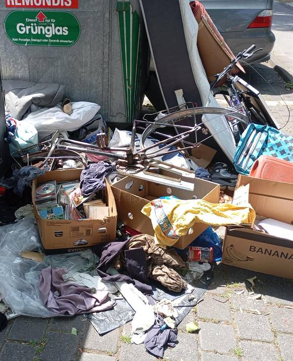 Vor Altkleider- und Glascontainern liegen Wäschestücke, eine Kiste mit CDs und Fahrräder ohne Reifen rum.