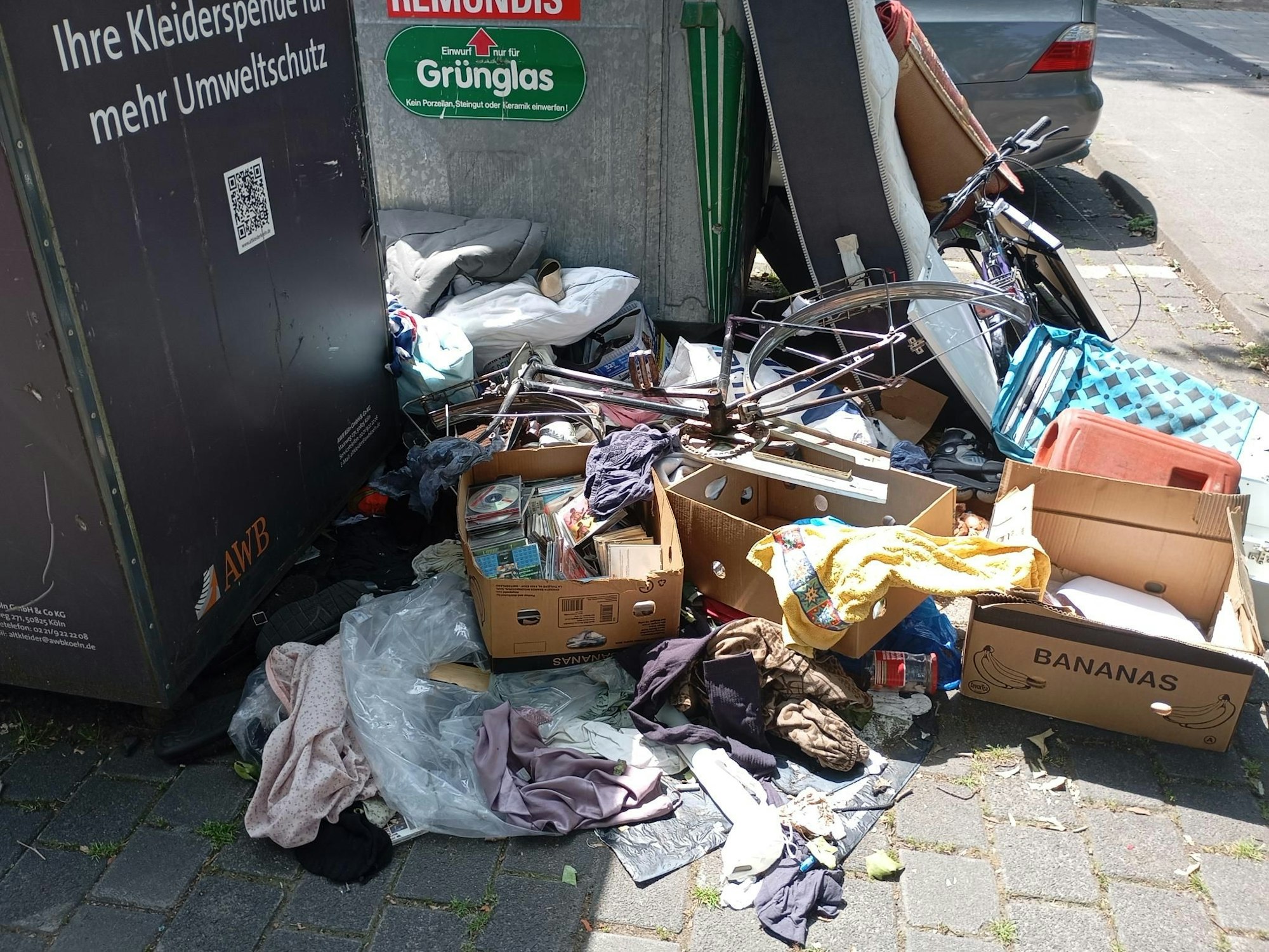 Vor Altkleider- und Glascontainern liegen Wäschestücke, eine Kiste mit CDs und Fahrräder ohne Reifen rum.