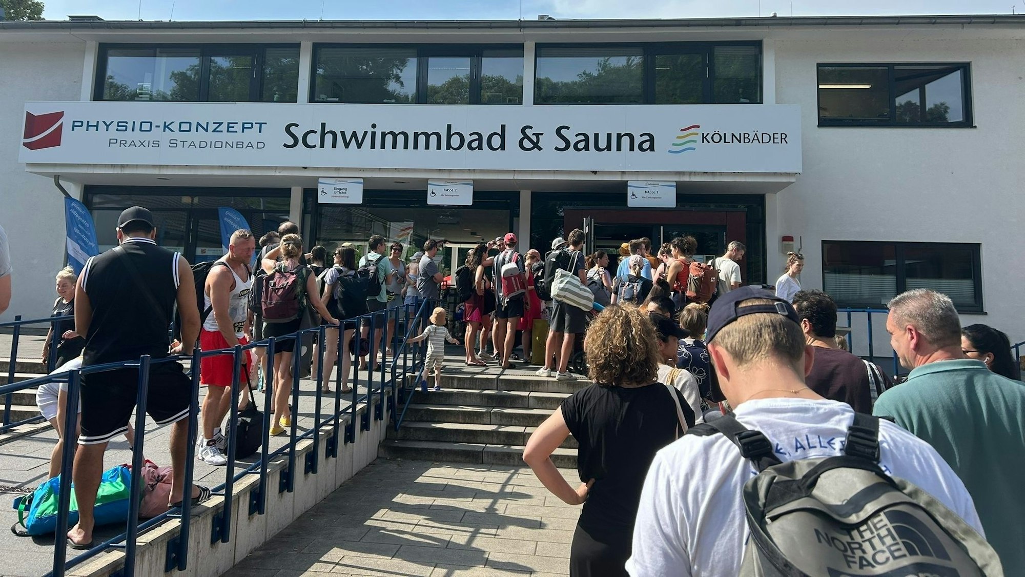 Vor einem Schwimmbad warten zahlreiche Menschen.