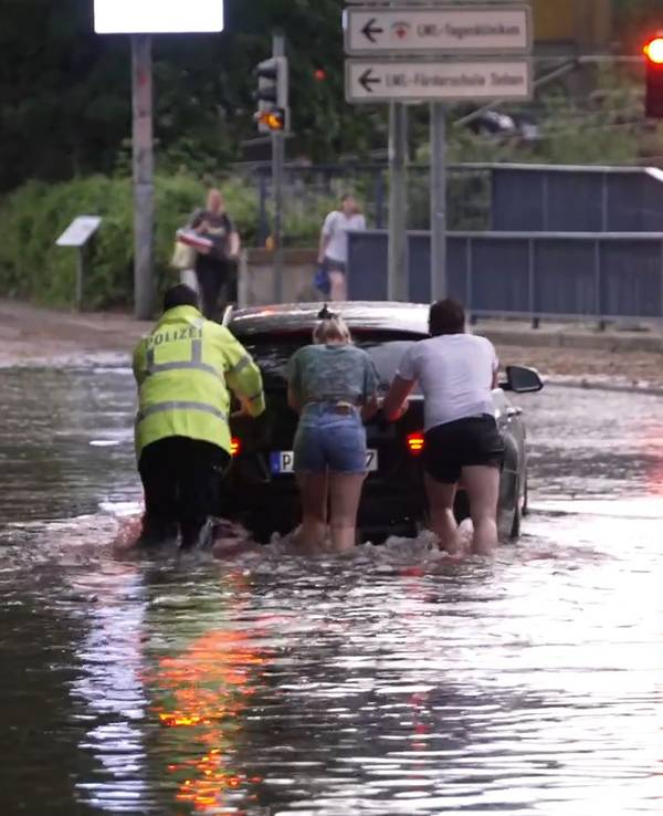 Menschen schieben ein Auto durch eine überflutete Straße. Ein schweres Unwetter hat am frühen Samstagabend große Teile des Kreisgebiets Paderborn getroffen.