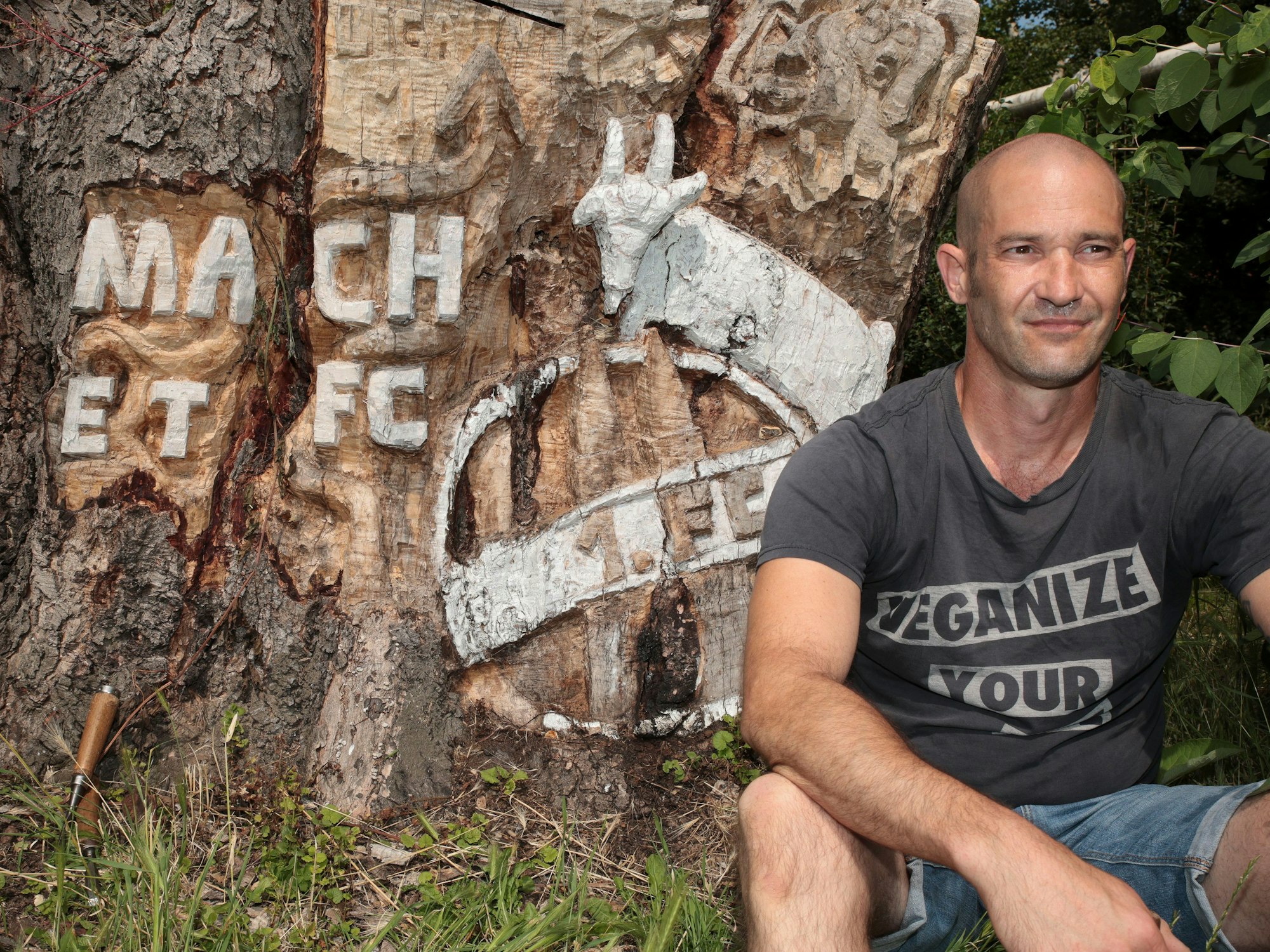 Ein Mann sitzt vor einem Baumstumpf, in den „Mach et FC“ geschnitzt wurde. Außerdem das FC-Wappen mit dem Geißbock.