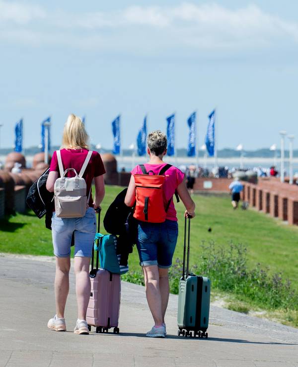 Zwei Frauen gehen bei sonnigem Wetter mit ihren Koffern über die Strandpromenade am Nordstrand der Insel Norderney.