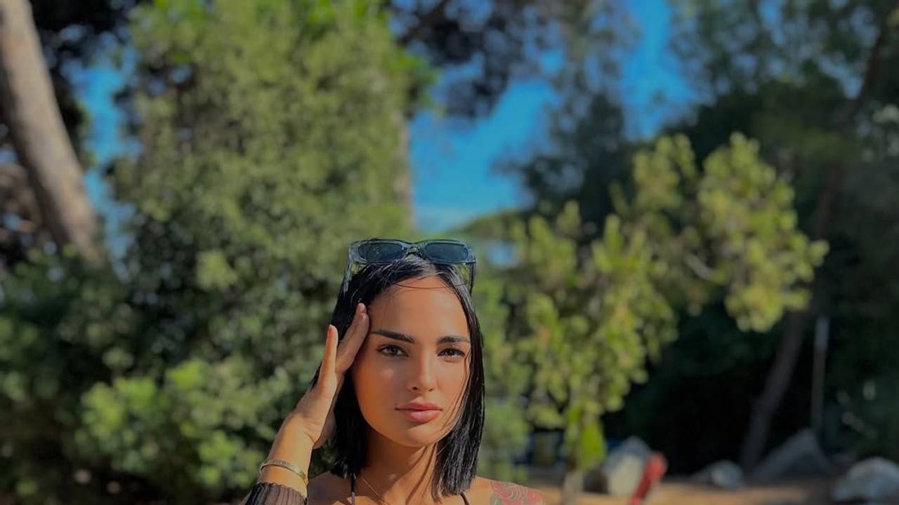 Claudia Bavel steht in einem bunten Bikini am Strand und fasst sich an den Kopf.
