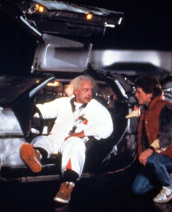 Die Darsteller von Doc Brown und Marty McFly posieren an einem DeLorean mit geöffneten Flügeltüren.