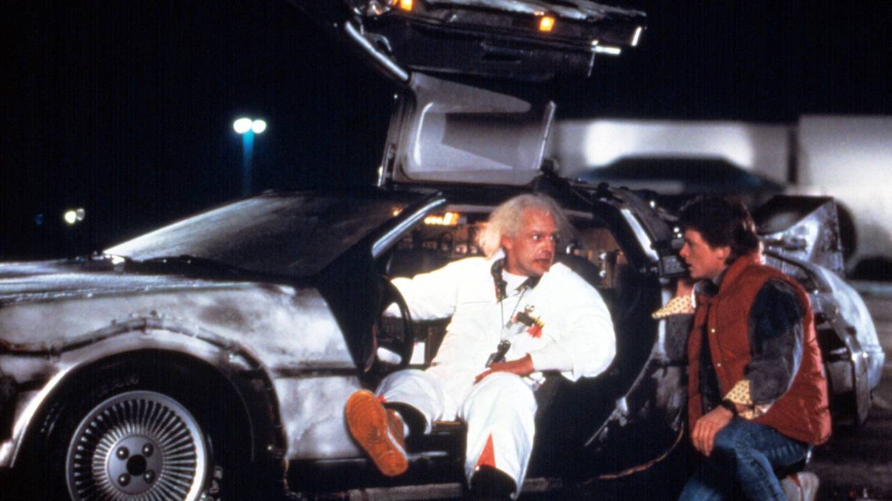 Die Darsteller von Doc Brown und Marty McFly posieren an einem DeLorean mit geöffneten Flügeltüren.