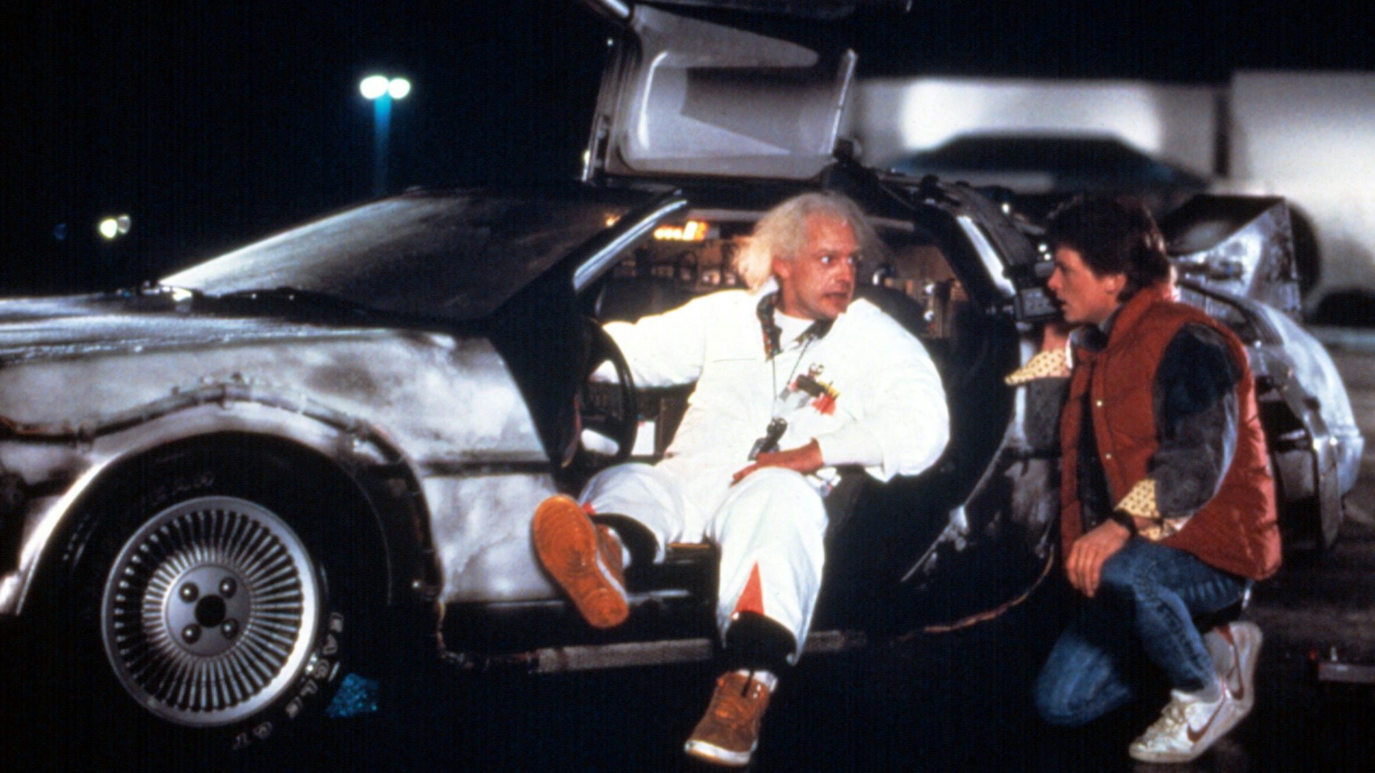 Die Darsteller von Doc Brown und Marty McFly posieren an einem DeLorean mit geöffneten Flügeltüren.