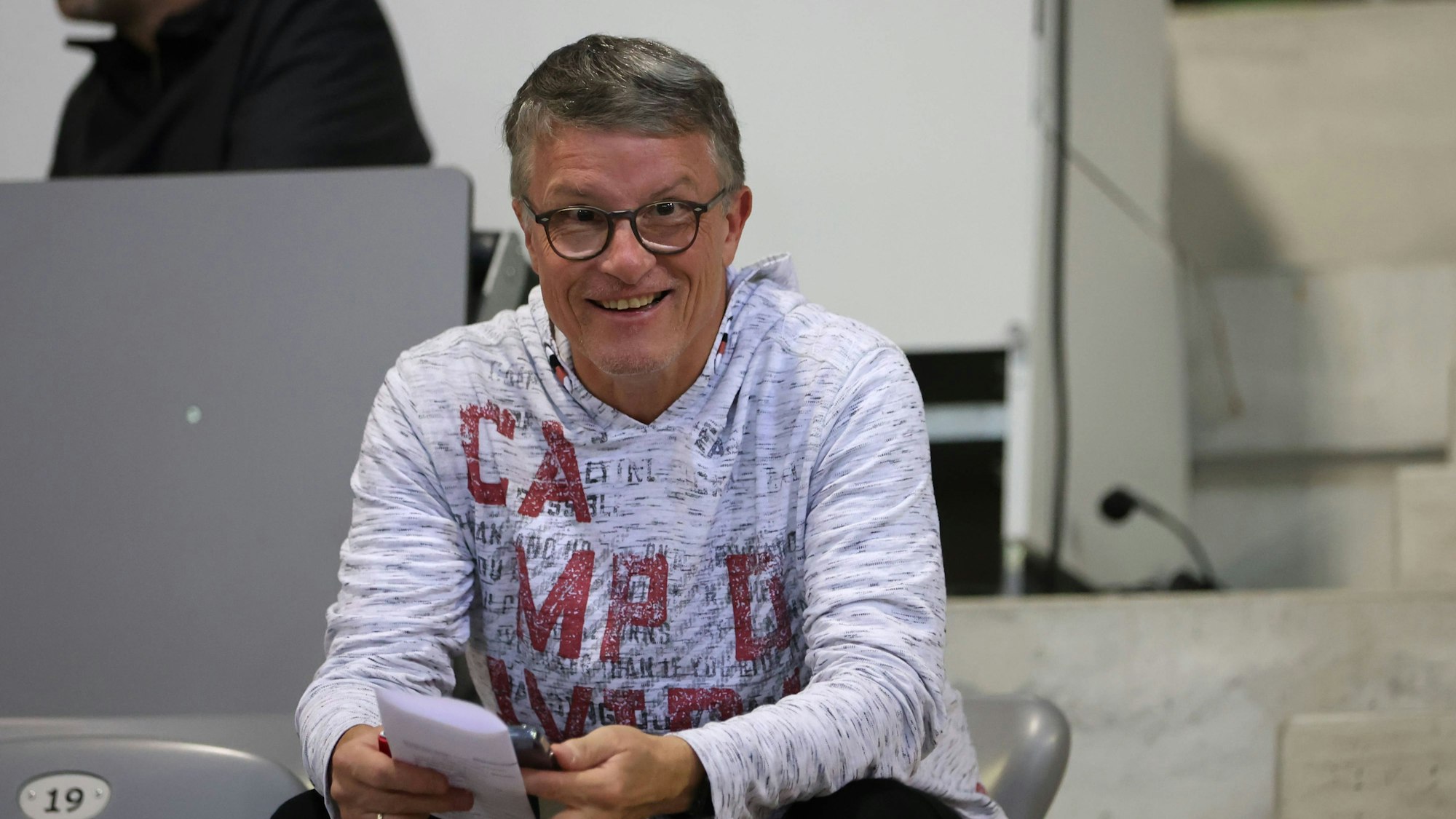 Sportreporter Bernd Schmelzer sitzt im grauen Hoodie auf einer Stadion-Tribüne.