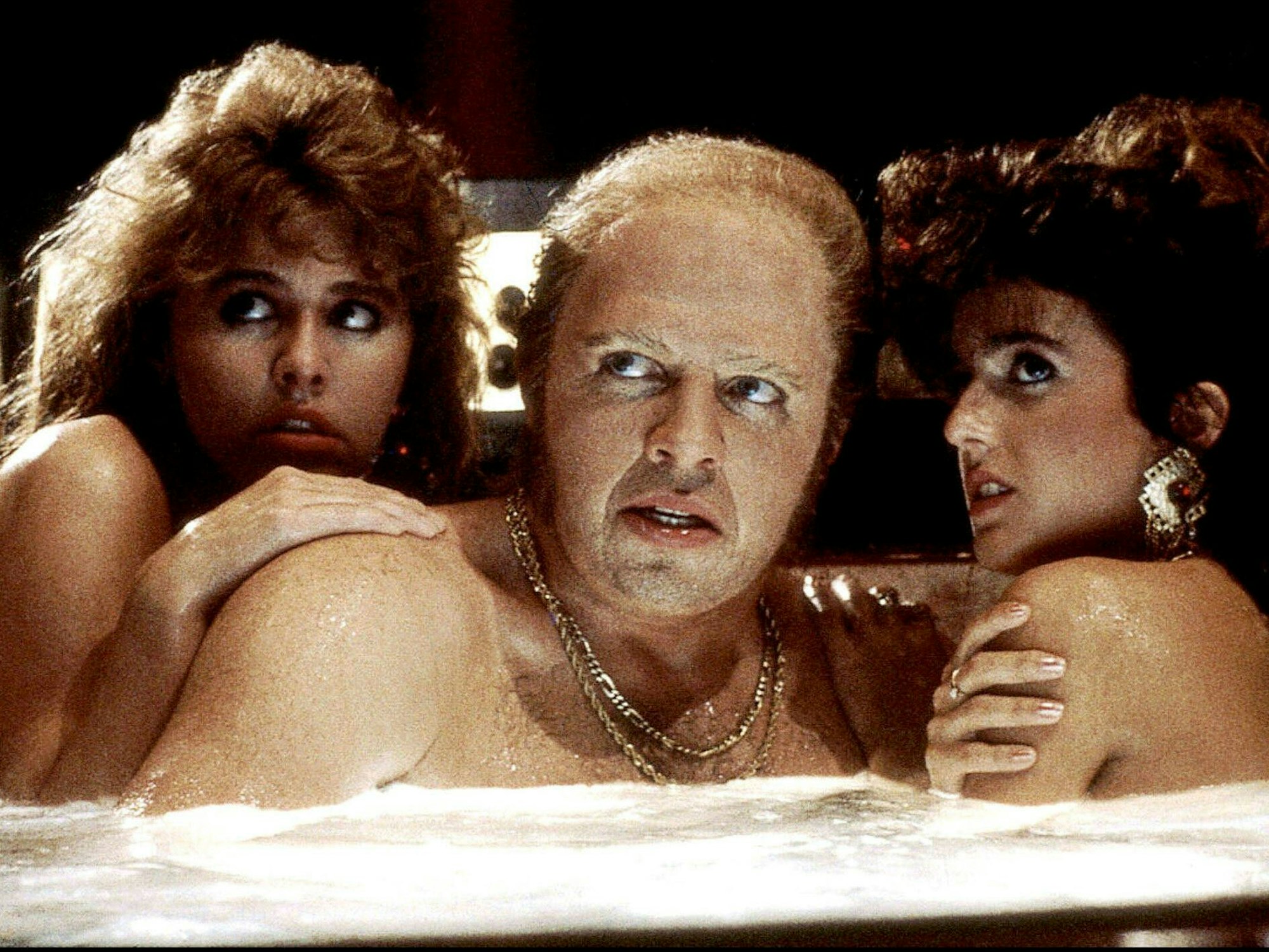Ein feister Mann mit Föhnfrisur und Goldketten sitzt mit zwei aufgedonnerten Damen in einem Whirlpool