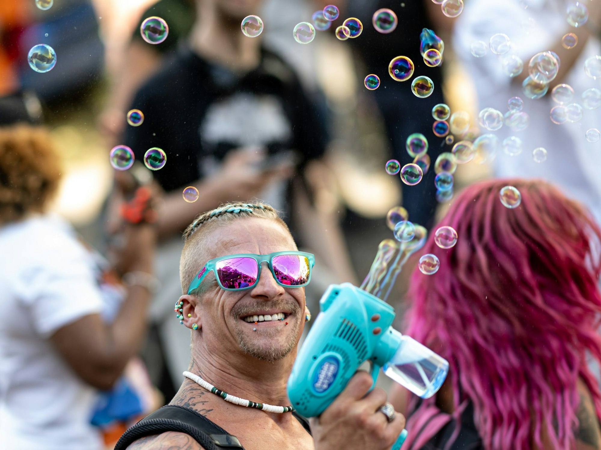 Eine Person mit Sonnenbrille und Piercings schießt mit einer kleinen Plastikpistole Seifenblasen in die Luft.