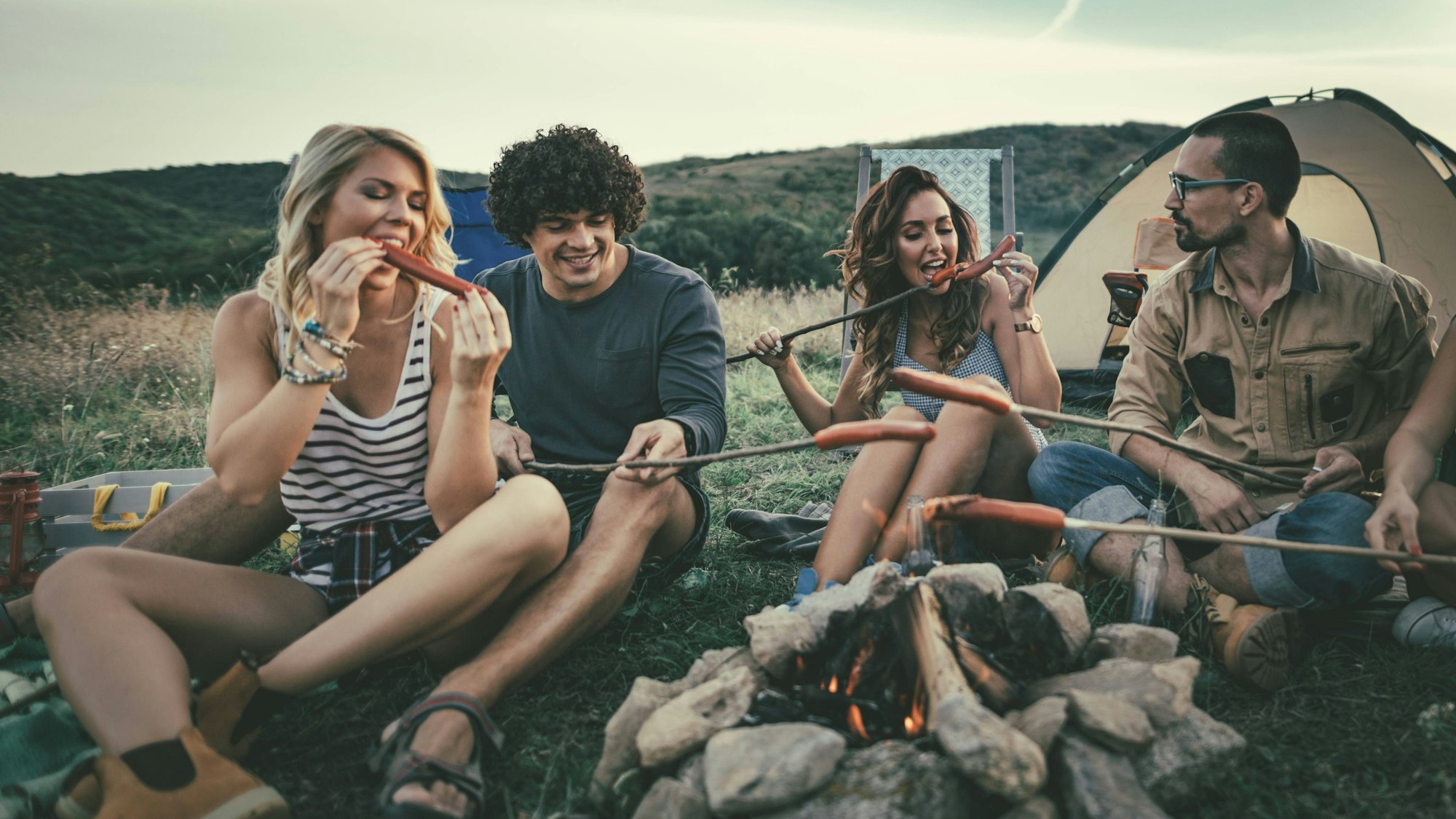 Junge Leute grillen Würstchen über einem Lagerfeuer und freuen sich.