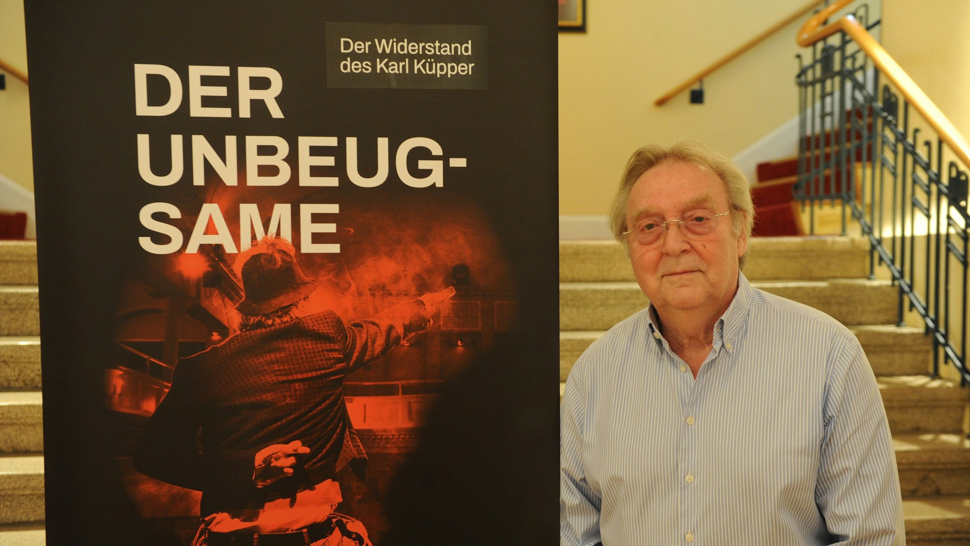 Gerhard Küpper steht neben dem Plakat des Theaterstücks über seinen Vater.