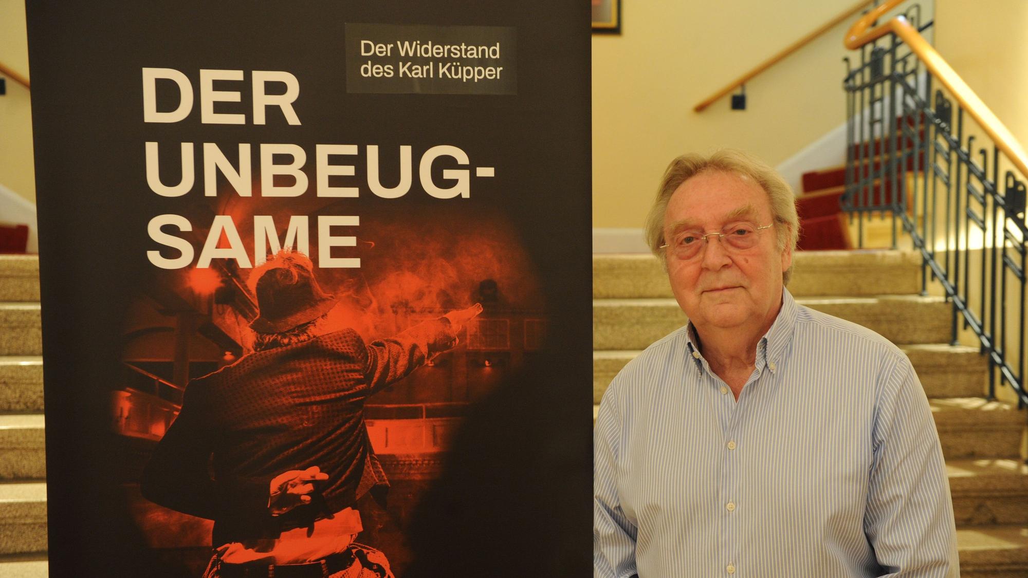 Gerhard Küpper steht neben dem Plakat des Theaterstücks über seinen Vater.