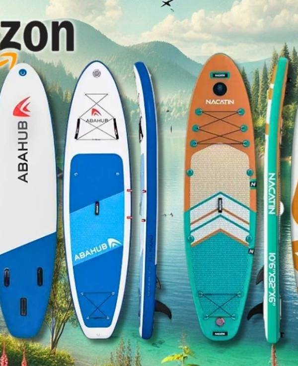 Eine farbenfrohe Auswahl an aufblasbaren Stand Up Paddling Boards aufgereiht vor einem idyllischen Seehintergrund mit Bergen und Wald.