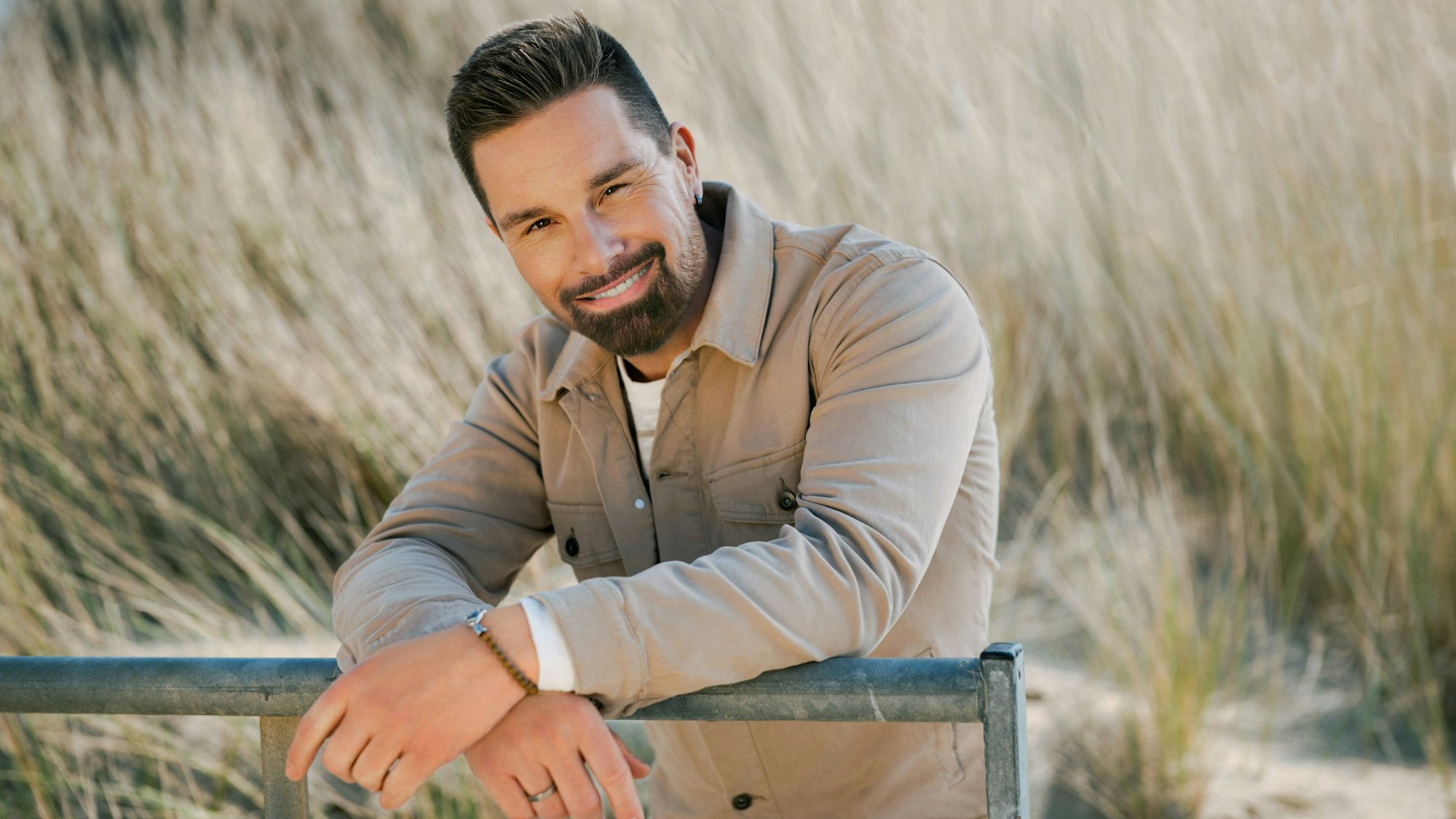 Ein Mann sitzt vor einer Dünenlandschaft und lächelt.
