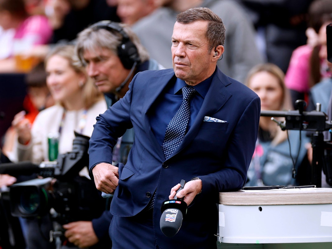 Lothar Matthäus steht während einer Pause mit Mikro in der Hand ans Moderatoren-Pult gelehnt.