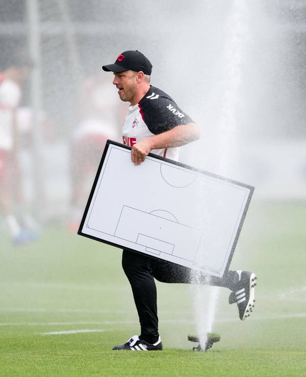 Köln-Trainer Lukas Kwasniok hält eine Taktiktafel in der Hand und läuft durch einen Rasensprenger.