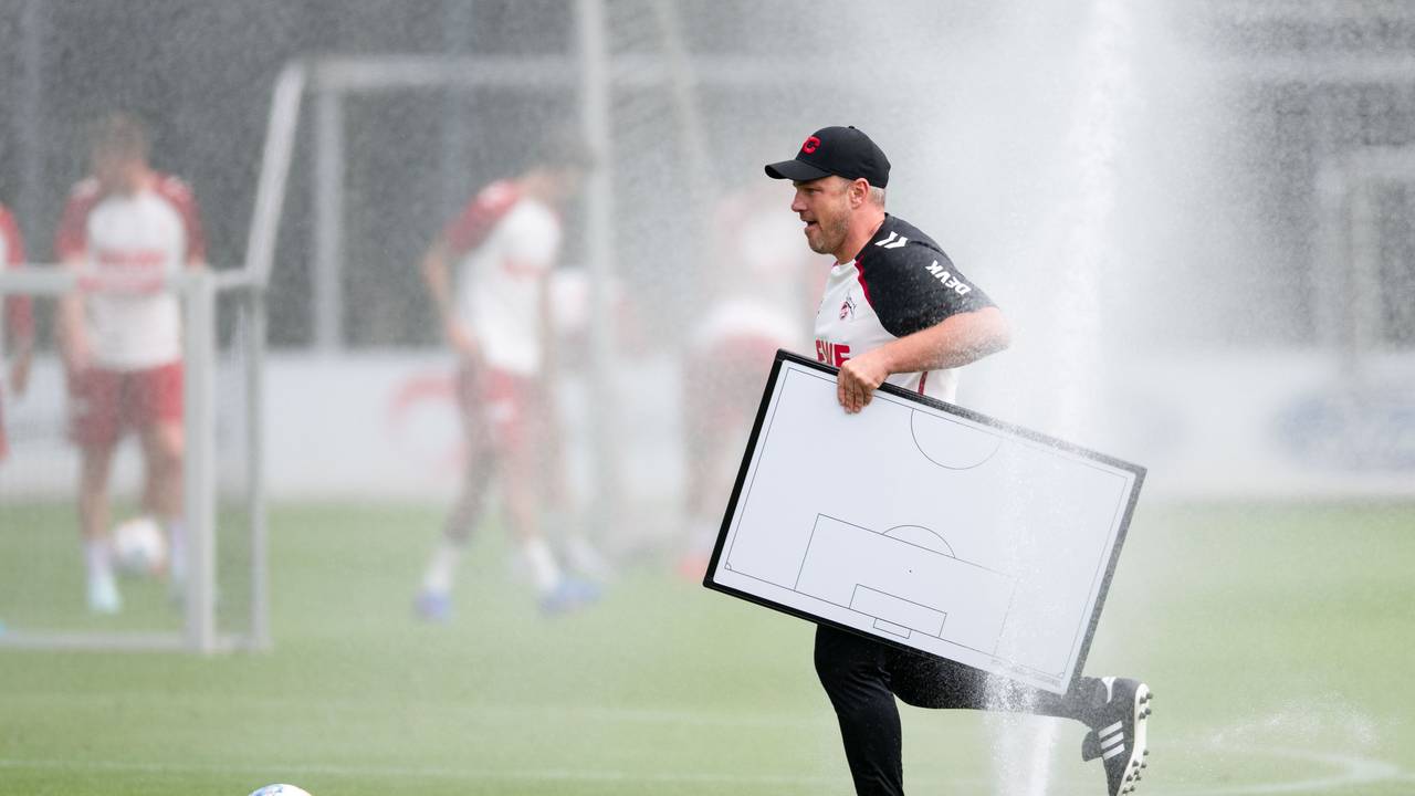 Köln-Trainer Lukas Kwasniok hält eine Taktiktafel in der Hand und läuft durch einen Rasensprenger.