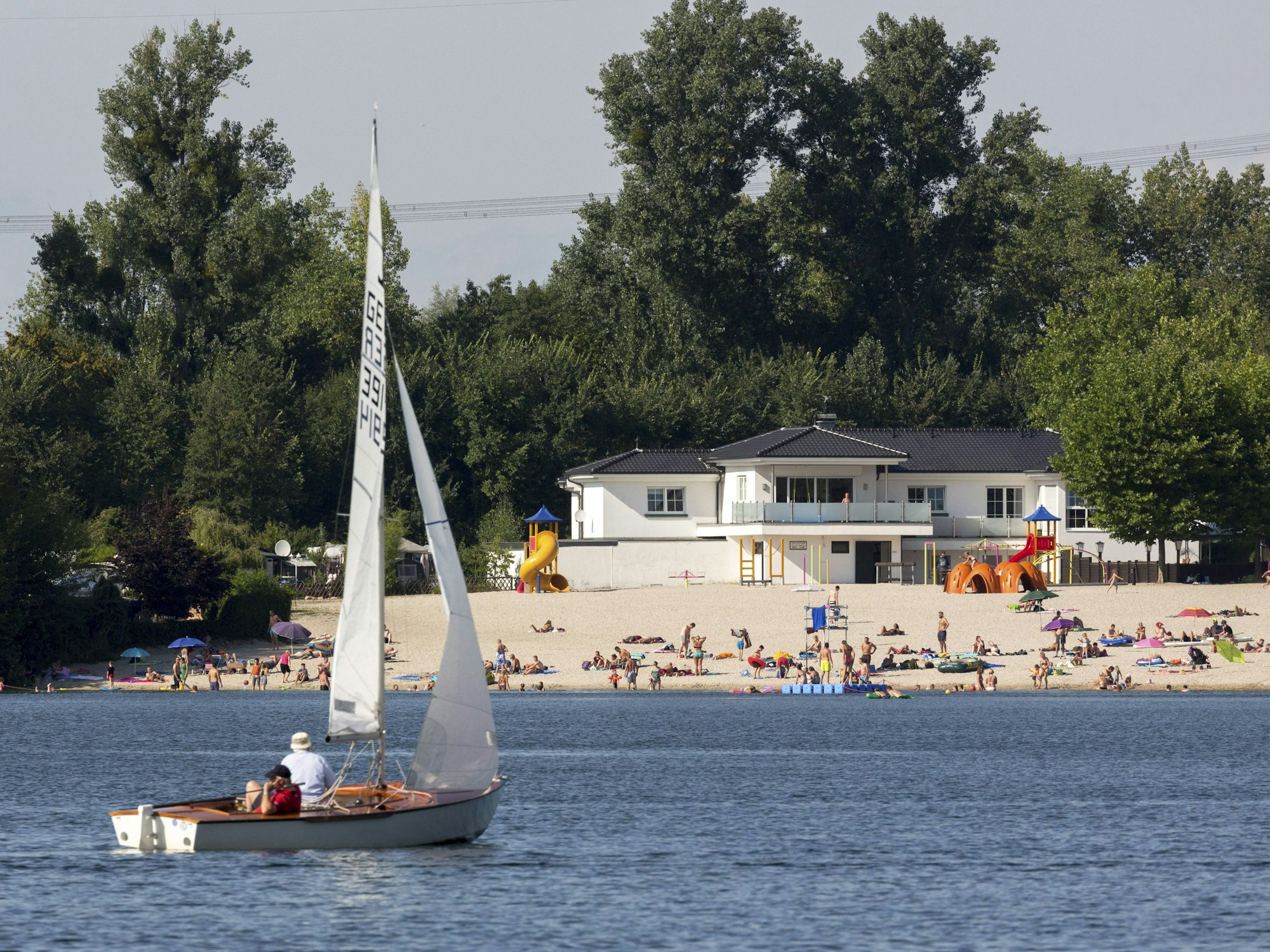 Ein Segelboot auf einem Badesee, im Hintergrund liegen Menschen am Sandstrand