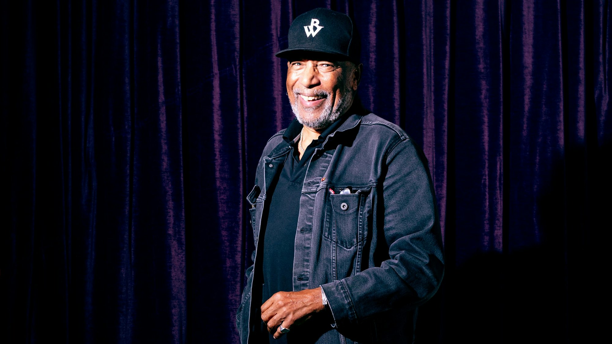 Ein Mann mit Baseballmütze steht vor einem dunklen Vorhang