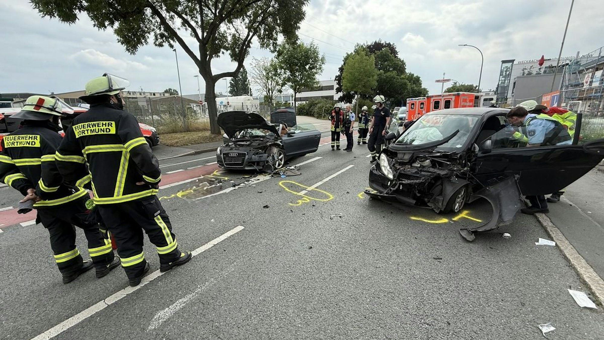 Zwei verunfallte Autos auf einer Straße, Feuerwehrleute im Hintergrund.