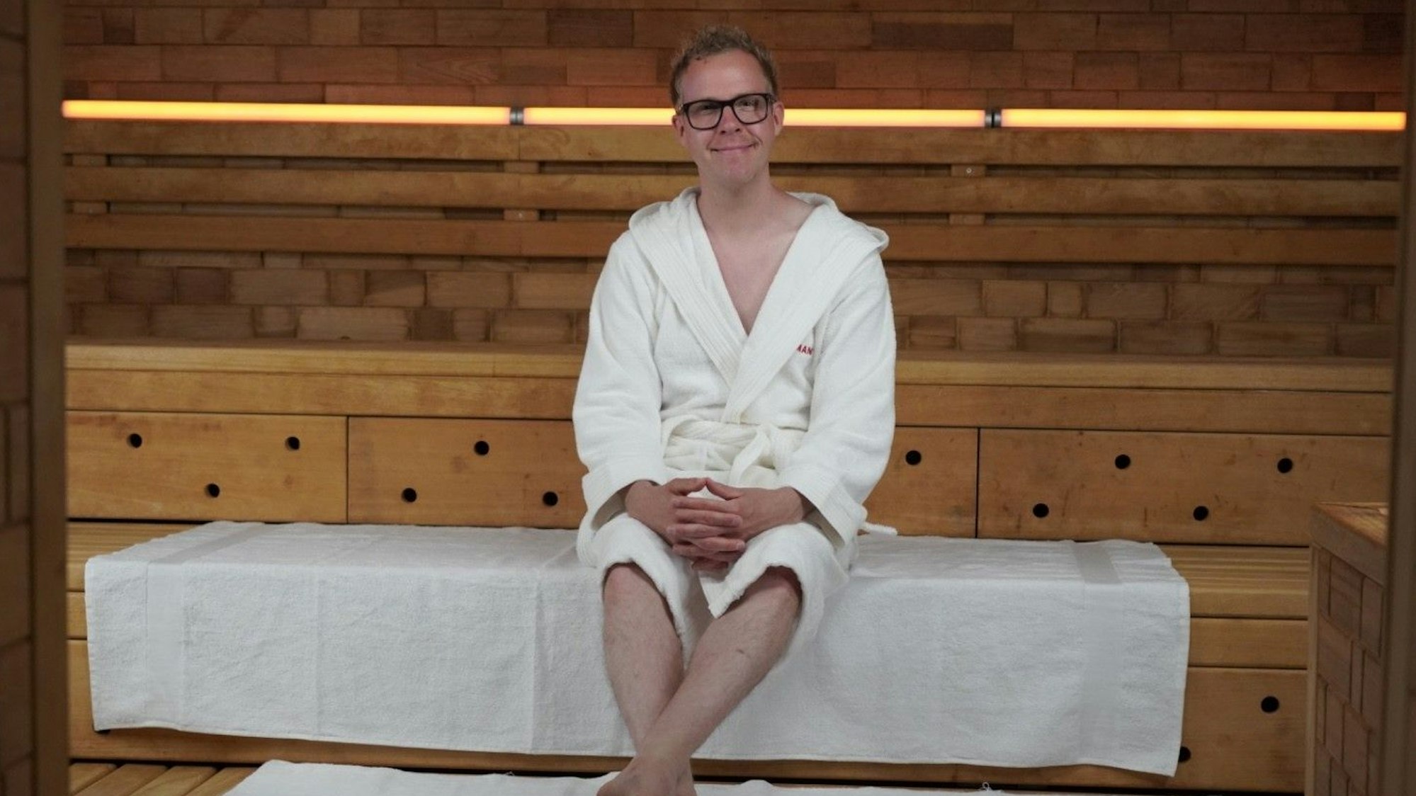 Ein Mann im weißen Bademantel sitzt auf der Holzstufe einer Sauna