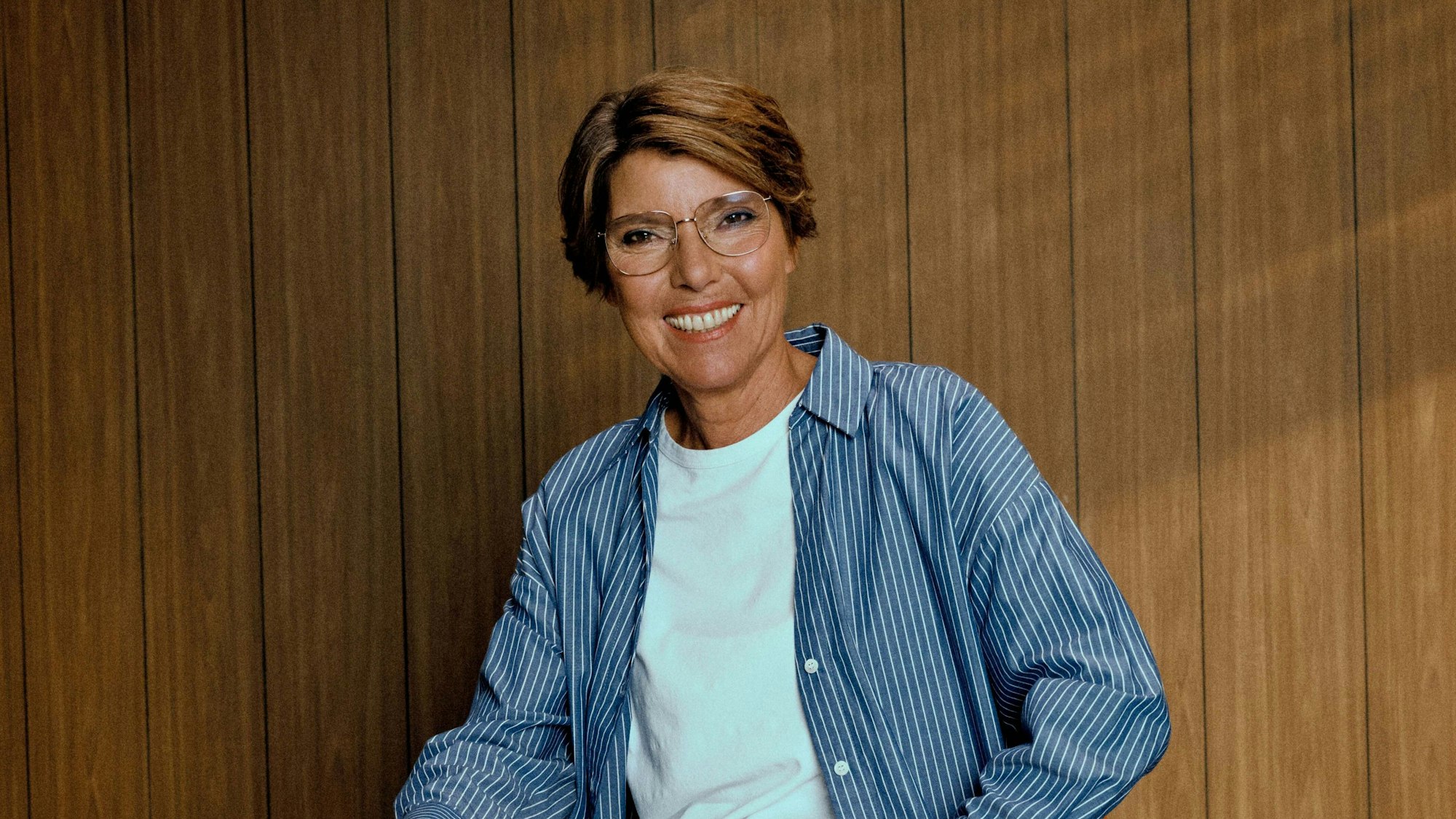Eine Frau im gestreiften Hemd sitzt vor einer Wand und lächelt