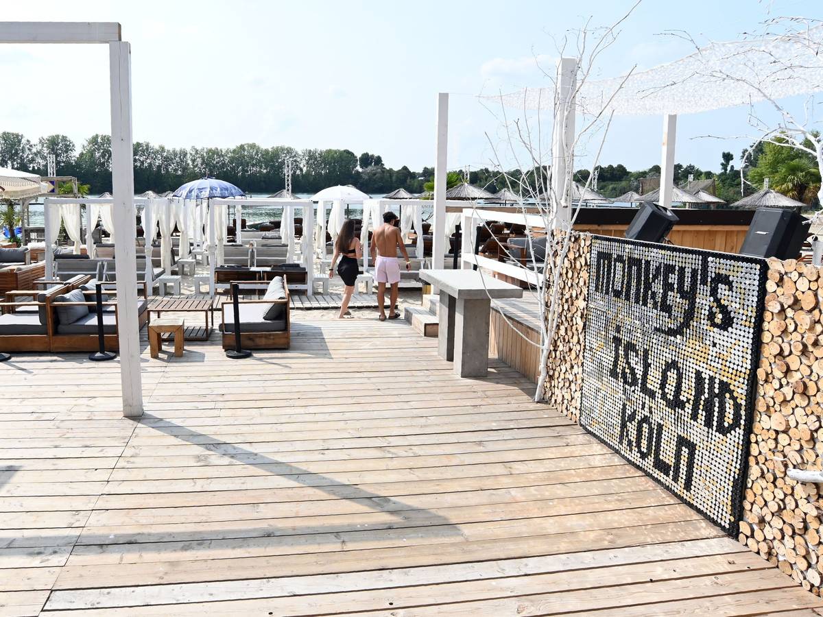 Am Beachclub Monkey's Island stehen weiße Schirme, ein Holzboden führt zum Strand.