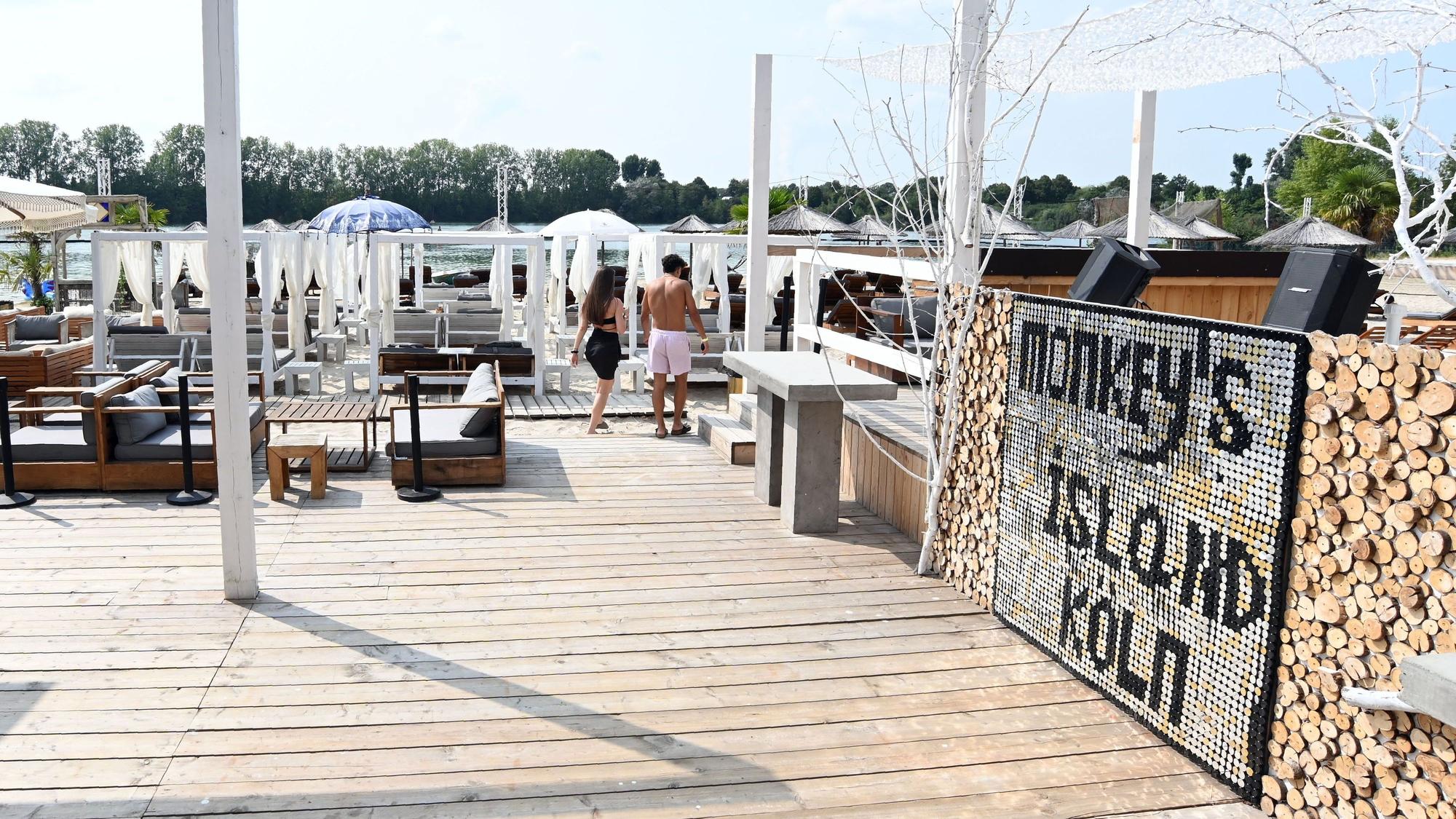 Am Beachclub Monkey's Island stehen weiße Schirme, ein Holzboden führt zum Strand.