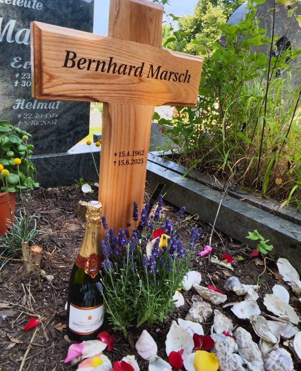 Vor einem Grabkreuz steht eine Flasche Crémant, liegen mehrere Austernschalen.