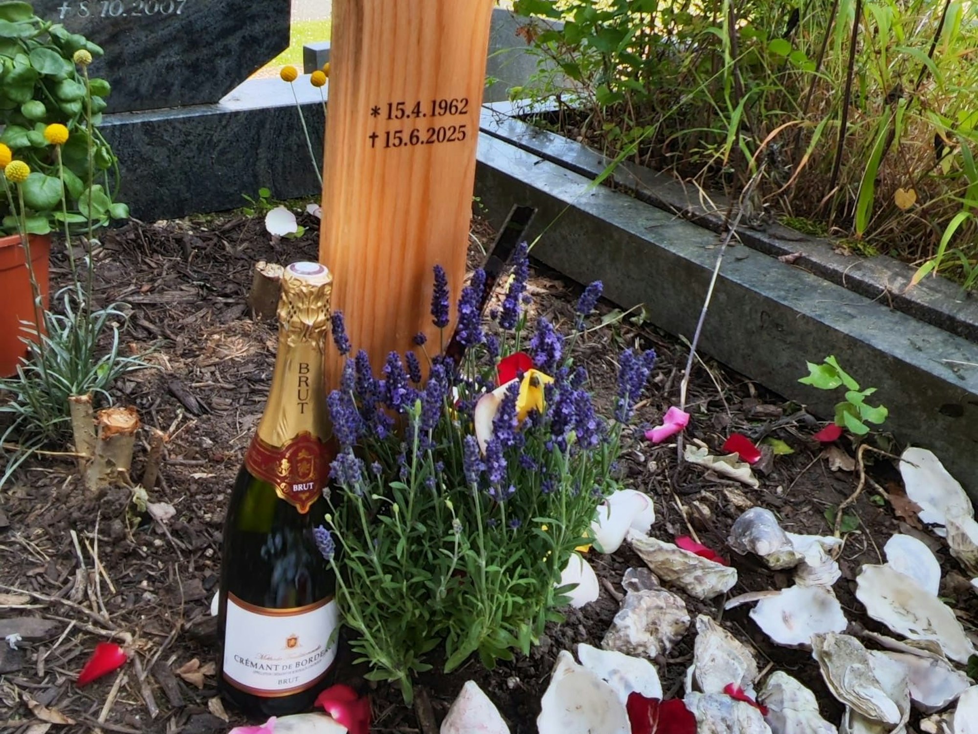 Vor einem Grabkreuz steht eine Flasche Crémant, liegen mehrere Austernschalen.