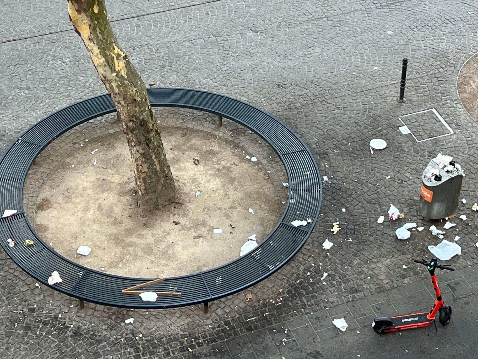 Auf einer runden Sitzbank liegt Müll, ebenso auf dem Boden rund um einen Mülleimer.